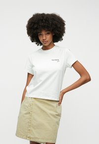 Camiseta blanca de manga corta con cuello redondo y logotipo negro en el lado izquierdo del pecho, combinada con una falda beige clara. Tela de algodón suave.