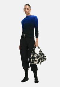 Blau-schwarzes, gemustertes Langarmoberteil, schwarze Hose mit Gürtel, Kampfstiefel, hält eine schwarz-weiß strukturierte Handtasche mit Trägern.