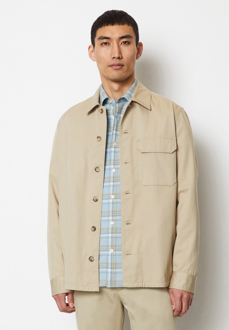 Beige lightweight jacket with a collar, five buttons, and a chest pocket, layered over a blue and white plaid shirt. Smooth fabric texture.