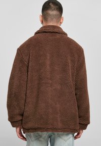 Brun, fluffy fleecejakke med krave og lange ærmer, der har et struktureret stof. Båret over lyseblå jeans, set fra ryggen.