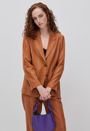 Femme aux cheveux roux bouclés portant un costume marron à rayures fines et tenant un sac à main violet sur un fond gris clair uni.