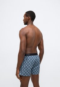 Γκρι μπόξερ σορτς με επαναλαμβανόμενο μοτίβο "HUGO" σε ανοιχτότερες και σκουρότερες αποχρώσεις, με μαύρη ελαστική μέση που έχει το λογότυπο.