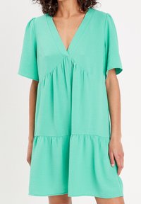 Une robe à manches courtes et décolleté en V de couleur vert menthe vif. Elle présente une taille froncée avec un design en couches et un tissu doux et texturé.