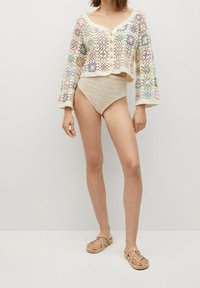 Gilet au crochet en crème avec des motifs floraux multicolores, coupe courte avec des boutons. Maillot de bain taille haute beige assorti et sandales.