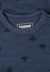 Marineblå bomulls T-shirt med ett subtilt biodlarmönster. Rund hals med tryckknappar, etiketten läser "hummel" i vitt. Mjuk textur.