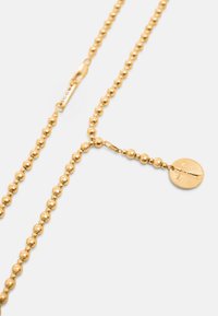 Collar de cadena con cuentas de color dorado que presenta un colgante medallón redondo con un diseño texturizado sobre un fondo claro y liso.