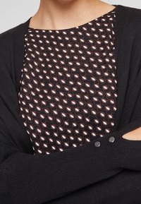Black cardigan with a ribbed texture, featuring a round neck and button detail. Underneath, a patterned top with a dark background and white accents.