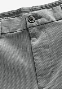Primer plano de una tela texturizada gris de unos pantalones con un cierre de botón y cinturilla. El material parece resistente con un patrón tejido.