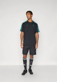 Juoda sportinė palaidinė su žaliomis juostelėmis, derančios juodos kelnės šortai ir juodos kelio aukščio kojinės su baltomis juostelėmis ir logotipo akcentais.