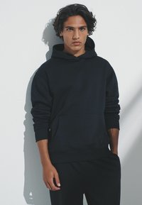 Tmavomodrá mikina s kapucňou z jemnej tkaniny s predným vreckom, rebrovanými manžetami a lemom; v páre s rovnakými nohavicami. Jednoduchý, moderný dizajn.