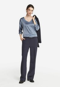 Navy-Breitbein-Hosen kombiniert mit einem hellblauen Satin-V-Ausschnitt-Top. Das Outfit umfasst einen schwarzen Blazer, der über dem Arm getragen wird, und schwarze Loafers.