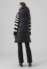 Gilet matelassé noir avec capuche, doté de manches rayées en crème et noir. Le gilet a une texture matelassée et une coupe longue jusqu'aux genoux.