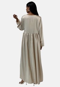 Lang, beige kjole lavet af glat stof med en rynket talje, lange ærmer og en rund halsudskæring, med et enkelt, flydende design.