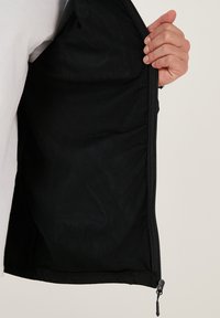 Interior de chaqueta de malla negra con textura suave, apertura con cremallera en la parte frontal y una mano agarrando la tela, mostrando su diseño ligero.