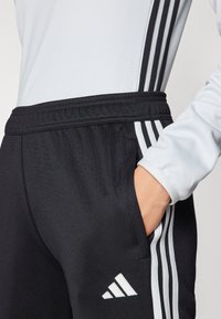 Černé sportovní kalhoty s bočními kapsami, elastickým pasem a bílým logem Adidas. Šedý dlouhý rukáv s černobílými pruhy na rukávech.