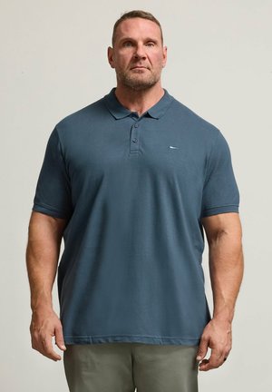 Muskelöser Mann mit kurzen Haaren, der ein dunkelblaues Poloshirt und beige Hosen trägt, steht vor einem schlichten hellen Hintergrund.