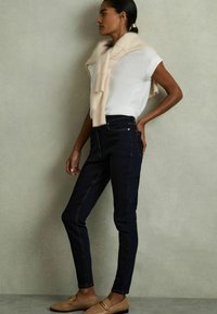 Mörkblå smala jeans med kontrasterande sömmar, kombinerade med en vit t-shirt och en ljusrosa stickad scarf hängande över axlarna, bärs med beige loafers.