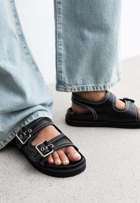 Svarta sandaler med två justerbara spännen, vit sömnad och en robust gummisula, tillsammans med ljusblå denimjeans.