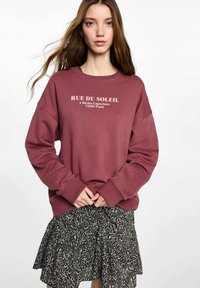 Felpa bordeaux con vestibilità oversize, polsini a coste e testo stampato. Abbinate a una gonna nera a fantasia, creando un look stratificato.