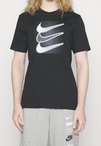 Černé tričko s krátkým rukávem, které má na čtvercovém grafickém potisku několik stylizovaných log Nike swoosh v bílé a šedé barvě. Materiál: bavlna.
