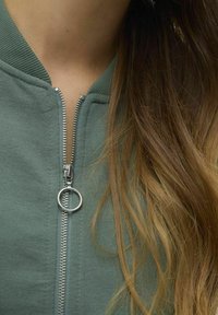 Vihreä pommitakki, jossa on ribbineuloskaulus, hopeinen vetoketju ja pyöreä vetoketjun vetimetarvike. Pehmeä, teksturoitu kangas, lyhyt malli.