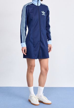 Persoon draagt een marineblauwe Adidas jurk met een ritssluiting en lichtblauwe strepen op de mouwen, witte sokken en witte sneakers, staande op een blauwe vloer.