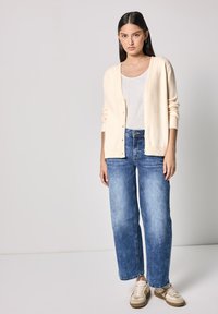 Cardigan lavorato a maglia crema con bottoni, indossato sopra una maglietta bianca e un paio di jeans blu larghi. Sneakers casual completano l'outfit.