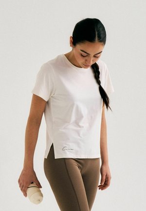 Femme avec une longue tresse foncée portant un t-shirt blanc à manches courtes et des leggings marron tenant une bouteille d'eau beige, regardant vers le bas.