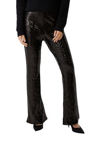Zwarte flonkerende flare broek met een glanzende textuur, strak rond de dijen en uitlopend bij de zoom; gecombineerd met zwarte schoenen.
