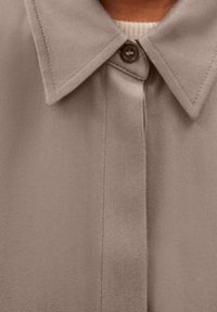 Ljusbrun skjorta med knäppning och spridd krage, som har ett texturerat tyg och en enda knappstängning i fronten.