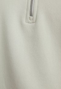 Primo piano di un tessuto beige chiaro con una cerniera argento corta e cucitura visibile al centro superiore.