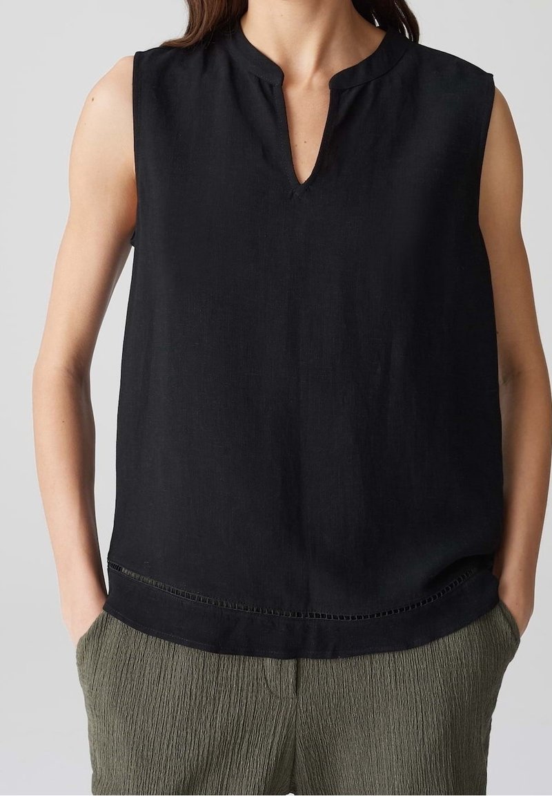 Haut noir sans manches avec encolure échancrée, porté avec un pantalon vert olive texturé, mains dans les poches, sur un fond clair uni.