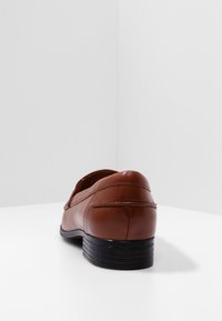 Mocassin en cuir marron avec un bout arrondi, un petit talon empilé et des détails de couture visibles sur les côtés, présenté sur un fond blanc.