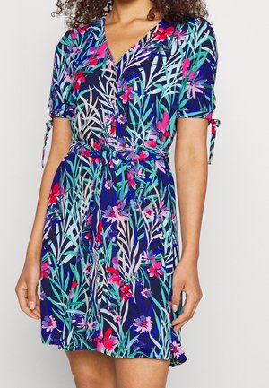 Vrouw draagt een knie-lange jurk met korte mouwen met een levendig bloemen- en bladerenpatroon in blauwtinten, groen, roze en rood.