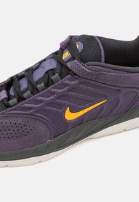 Lila sportcipő, texturált szövet felső részel, fekete hálós nyakkal, sárga Nike logóval és szürke gumitalppal. Laposszálas és perforált.
