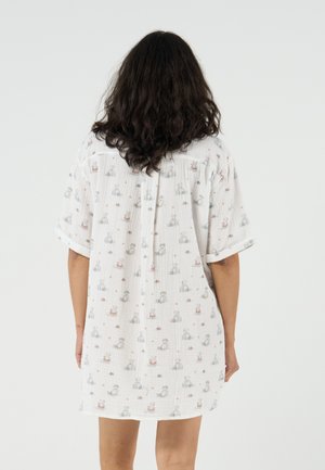 Personne aux cheveux longs et foncés portant une robe blanche ample avec un petit motif animalier, debout de dos sur un fond uni.