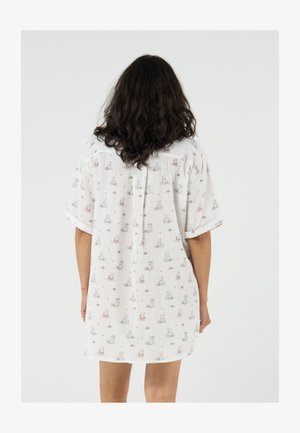 Personne aux cheveux longs et foncés portant une robe blanche ample avec un petit motif animalier, debout de dos sur un fond uni.