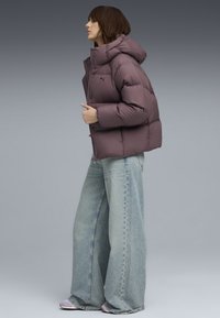 Lilla pufferjakke med hætte, quiltet design og forsyningsknapper, parret med løse lyseblå jeans, set fra siden.