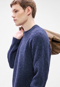 Ναυτικό πλεκτό πουλόβερ με υφή και στρογγυλή λαιμόκοψη, φορεμένο από μοντέλο που κρατάει μια καφέ τσάντα στον ώμο του.