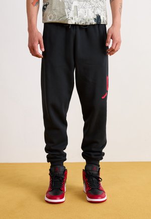 Mann in schwarzen Jogginghosen mit rotem Logo auf dem Oberschenkel und rot-schwarz-weißen High-Top-Sneakern, der auf einem gelben Boden steht.