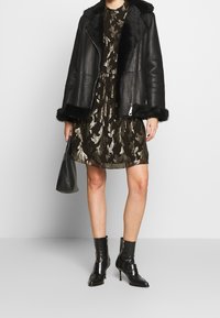 Veste en cuir noir avec une bordure en fausse fourrure, superposée à une robe sombre avec un motif floral doré ; assortie à des bottines noires et un sac coordonné.