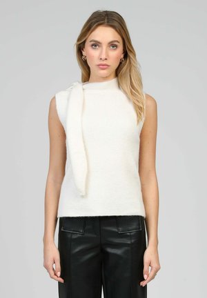 Femme aux longs cheveux blonds portant un pull sans manches blanc avec un détail d'écharpe et un pantalon en cuir noir, debout devant un fond gris.