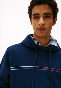 Mørkeblå hettegenser med hvite og røde striper og "TOMMY JEANS"-logo. Den har en hette med snor og et lagdelt rutete skjorte under.