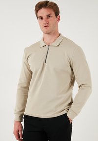 Polo shirt beige a maniche lunghe con colletto zip, polsini a coste e un tessuto testurizzato, abbinato a pantaloni neri.