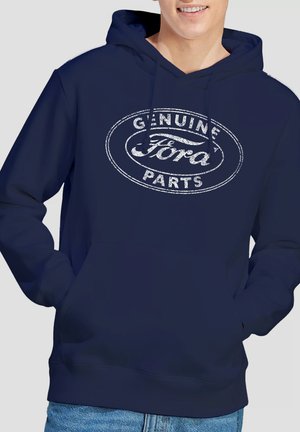 Man wearing navy blue hoodie with "Genuine Ford Parts" printed in distressed white text, hands in front pocket, light blue jeans visible.