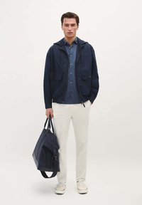 Marine lichtgewicht jack met twee voorzakken, blauwe overhemdd, witte broek en marine sporttas. Soepele stof en ontspannen pasvorm.