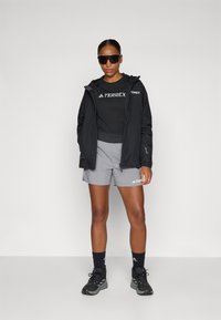 Svart lett jakke, grå shorts, svart skjorte med "TERREX"-logo, kombinert med svarte sportssko og solbriller.