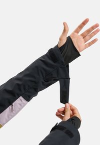 Schwarzer wasserdichter Jackenärmel mit einem Klettverschlussriemen, der lila und schwarze Farbblöcke aufweist und eine Reißverschlusstasche am Unterarm hat.