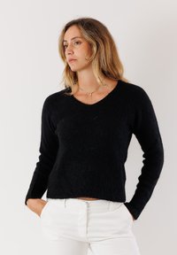 Maglione nero, con texture, a V e maniche lunghe, caratterizzato da una vestibilità comoda e un orlo cropped. Indossato sopra pantaloni bianchi.