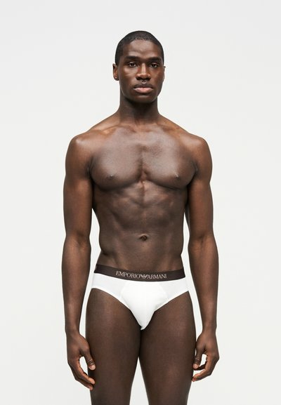 Μυώδης άντρας σε όρθια στάση, φορώντας λευκά εσώρουχα Emporio Armani με μαύρη ζώνη μέσης, μπροστά σε απλό φωτεινό φόντο.